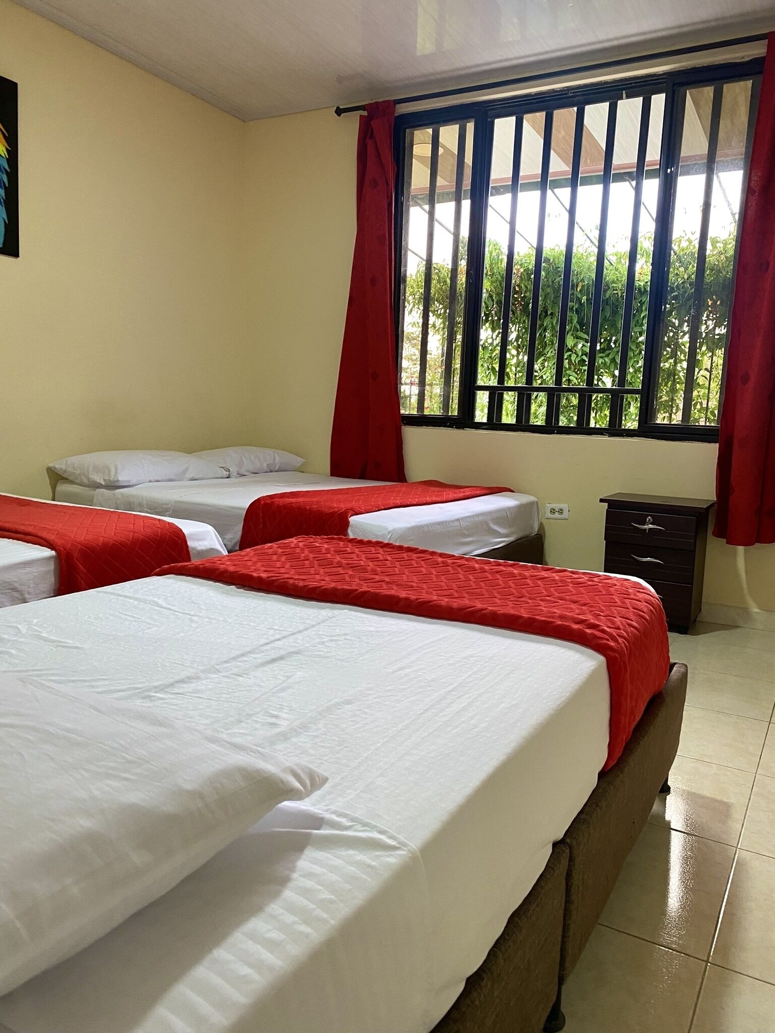 A hotel room with two beds, each with white linens and red bedspreads, a window with bars, and a view of greenery outside.