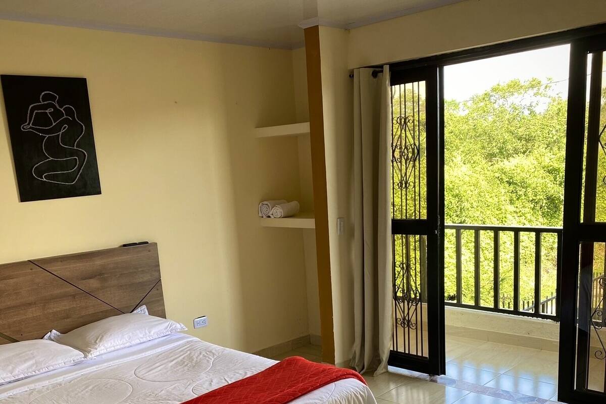 Ein Schlafzimmer mit einem Bett, einem Kopfteil, einer roten Decke, Wandkunst und einem Balkon mit Blick auf Bäume.