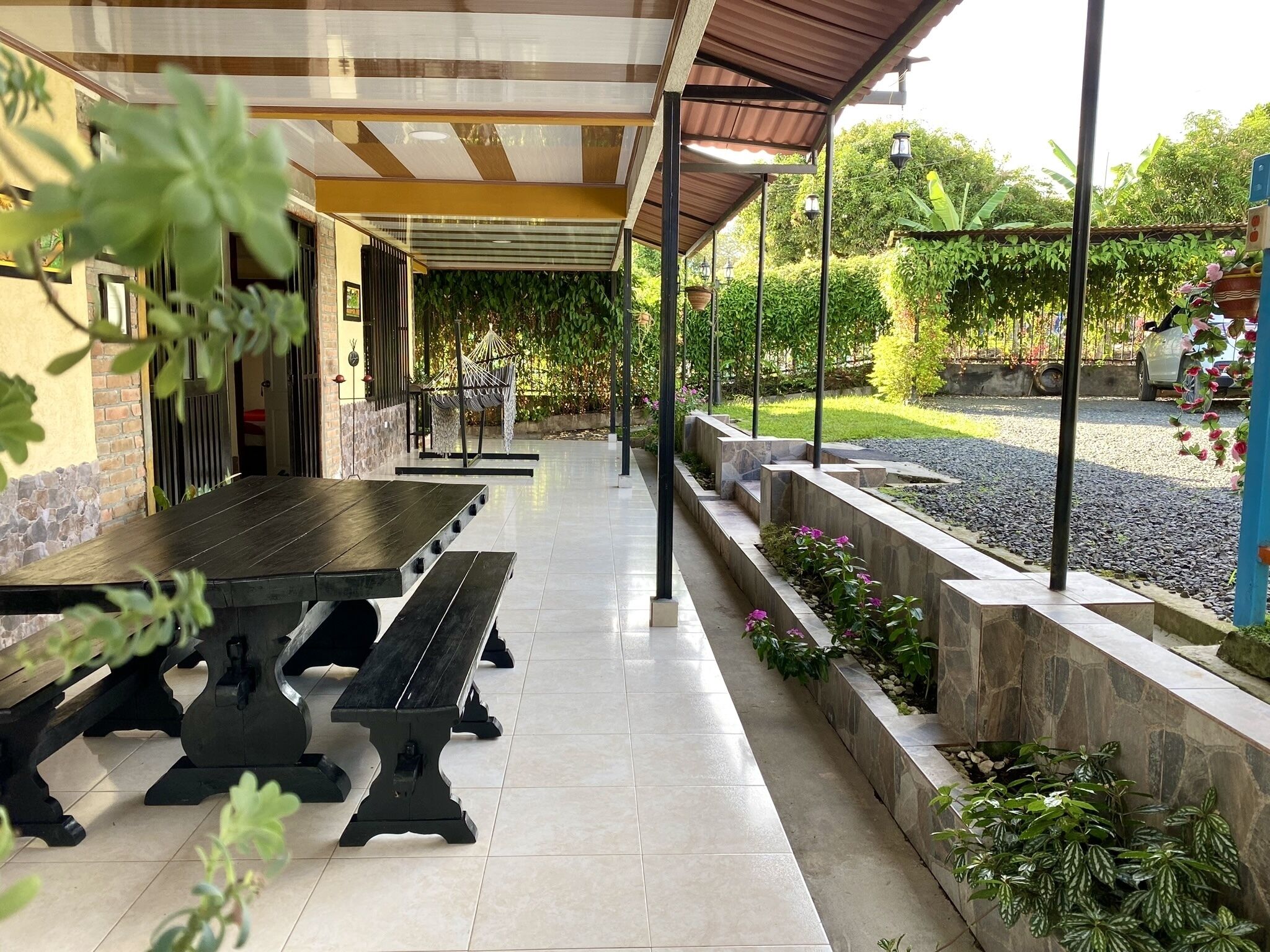 A covered outdoor area with a long table and benches, surrounded by stone planters and greenery.