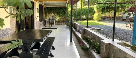 A covered outdoor area with a long table and benches, surrounded by stone planters and greenery.
