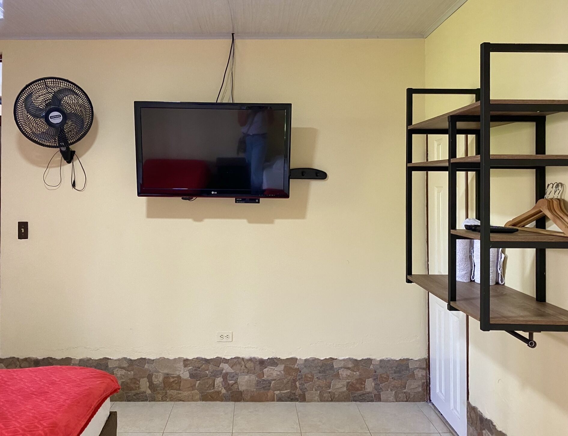 Una habitación con un televisor de pared, un ventilador de pie y un estante de madera.