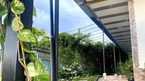 Uma área externa coberta com uma escadaria de pedra que leva a uma entrada coberta, cercada por vegetação e um portão de metal.