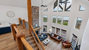 Amplia sala de estar con chimenea de piedra, escalera de madera y grandes ventanales con vistas al paisaje circundante.