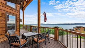 Terraza de madera con mesa y sillas, con vista a un lago y una cadena montañosa distante.