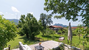 Eine Holzdielen-Terrasse mit Gartenmöbeln, einem Tisch mit Blumen und Blick auf ein Haus und Berge im Hintergrund.