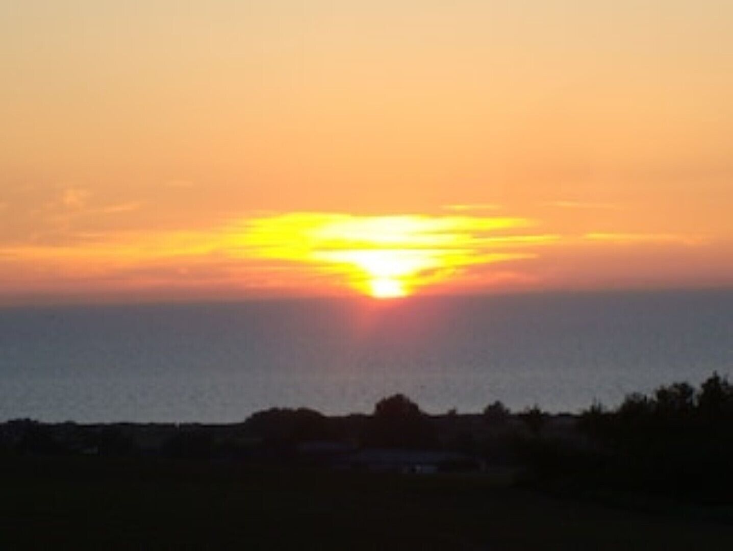 Un coucher de soleil sur une étendue d’eau, avec en silhouette au premier plan un paysage et un bâtiment.