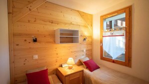 A small, cozy room with wooden walls, two beds, a nightstand with a lamp, and a window with a curtain. - 5m from ski slopes, 4th floor, view mountain, balcony, ski locker, 25m², Valmorel (Valmorel)