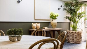 A dining area with a round wooden table, wicker chairs, potted plants, and a large window providing natural light.