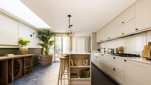 A modern kitchen with a central island, wooden stools, a bookshelf, and a large window allowing natural light.