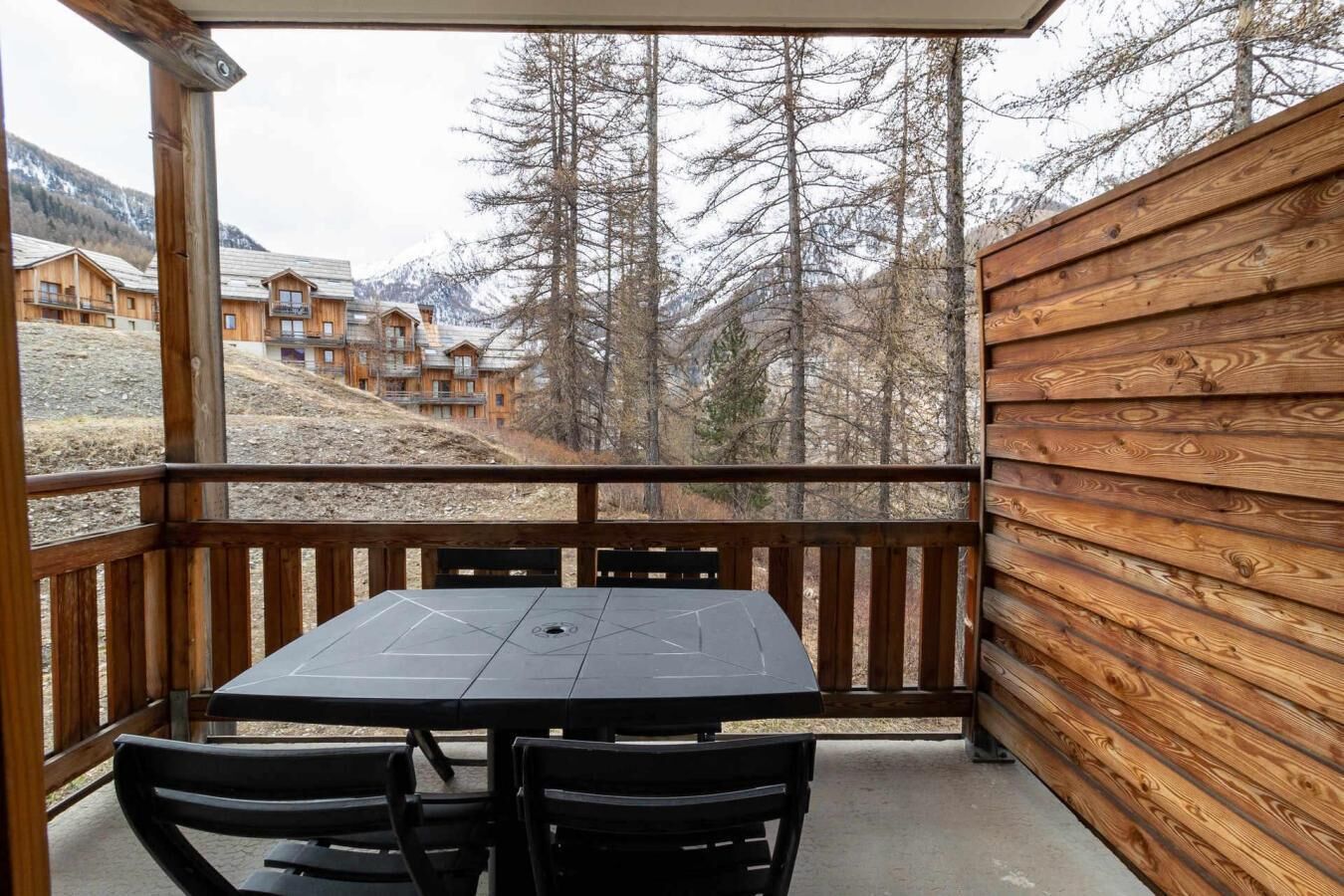 Une terrasse en bois avec une table et des chaises, offrant une vue sur une région montagneuse où l’on aperçoit des bâtiments au loin.