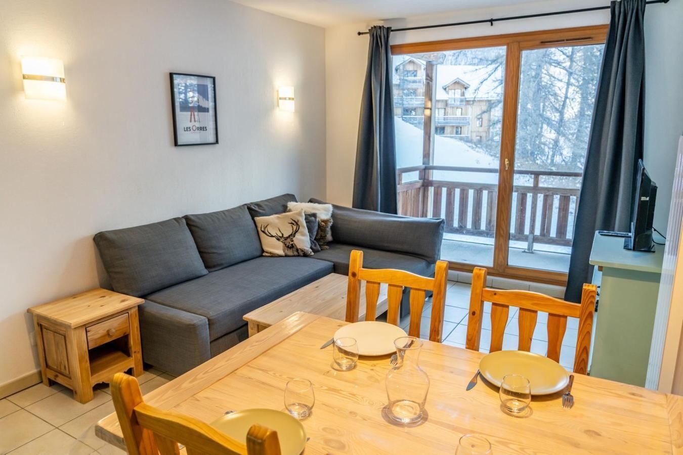Un salón acogedor con un sofá gris, una mesita de madera y una mesa de comedor preparada para dos personas, con copas de vino.