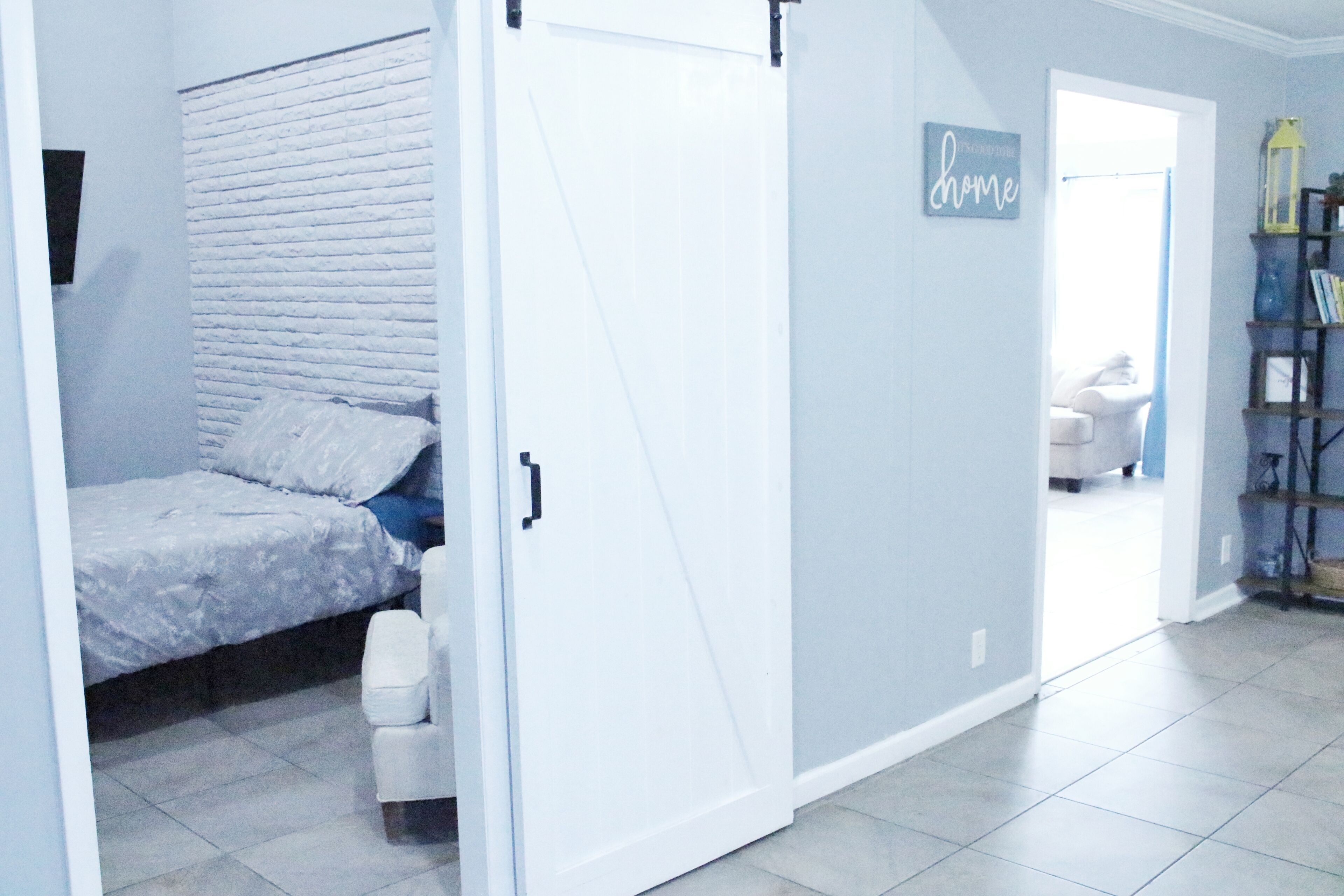 A room with a bed, a white door, a 'home' sign, a bookshelf, and a chair.