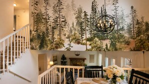 A dining area with a table set for two, a chandelier, and a wall with a forest mural. - Family Retreat | Treehouse + Game Room | Near Silverwood (Post Falls)