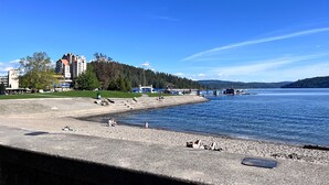 A waterfront area with a concrete walkway, a sandy beach, and buildings in the background. - Family Retreat | Treehouse + Game Room | Near Silverwood (Post Falls)