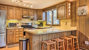 Una cocina con armarios de madera, un refrigerador de acero inoxidable, un microondas, un horno y un lavamanos. Hay tres taburetes de madera y un bote de basura.