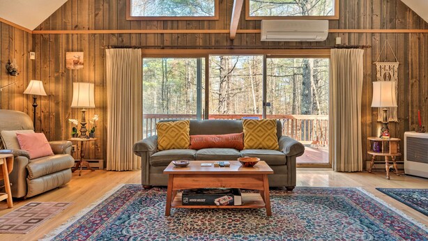 Una sala de estar acogedora con detalles de madera, un ventanal con vistas a árboles, un sofá con cojines, una mesa de centro y una lámpara.