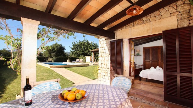 A covered outdoor seating area with a table set for two, a bottle of wine, and a bowl of fruit. - FONTXICA (JUANITO ). Nice and cosy villa (Puerto Pollensa)