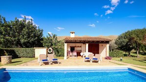 A swimming pool with lounge chairs and a patio area, surrounded by greenery and a building with a tiled roof. - FONTXICA (JUANITO ). Nice and cosy villa (Puerto Pollensa)