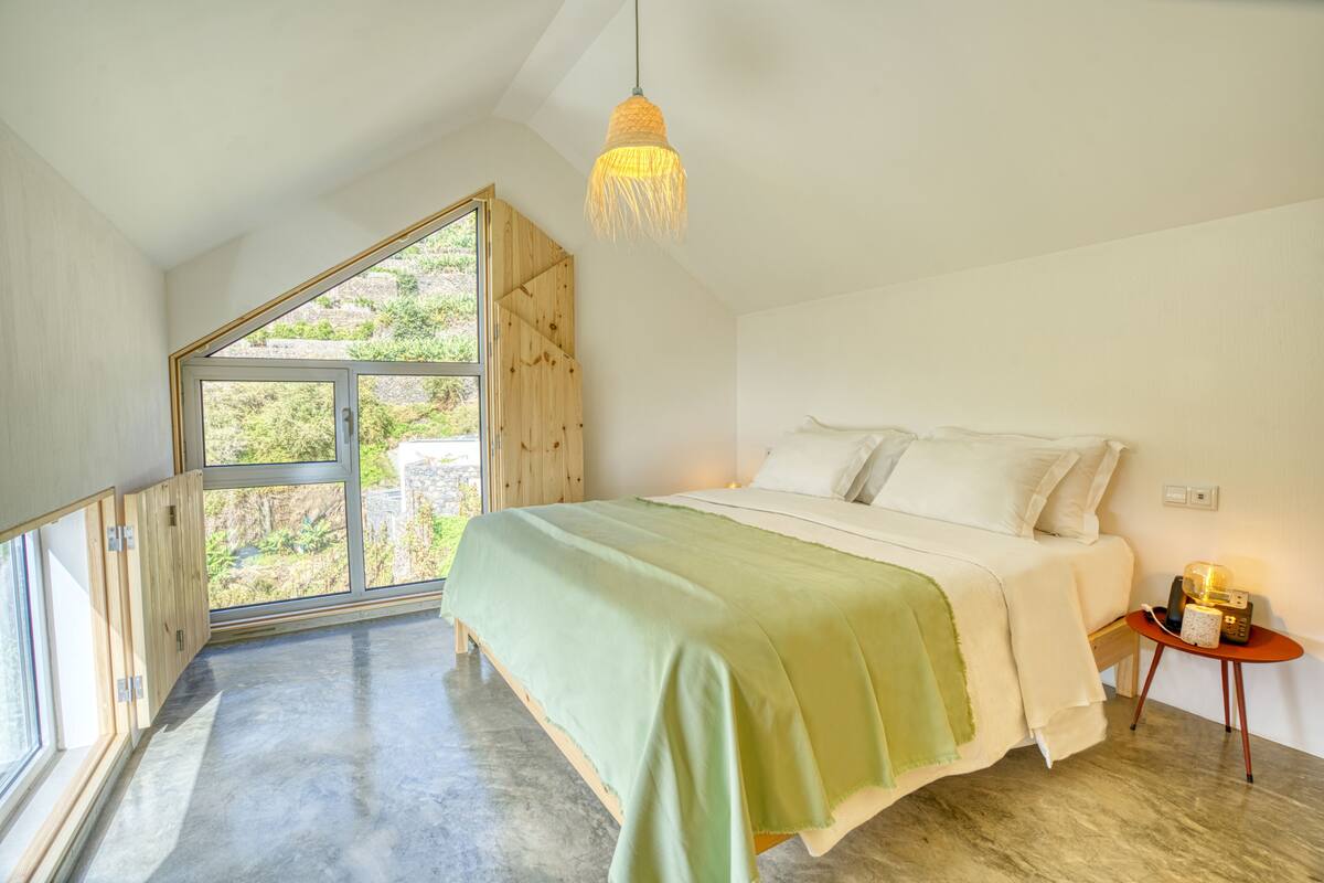 Quarto com uma janela grande, cama com manta verde, guarda-roupa de madeira e mesa de cabeceira pequena com abajur.