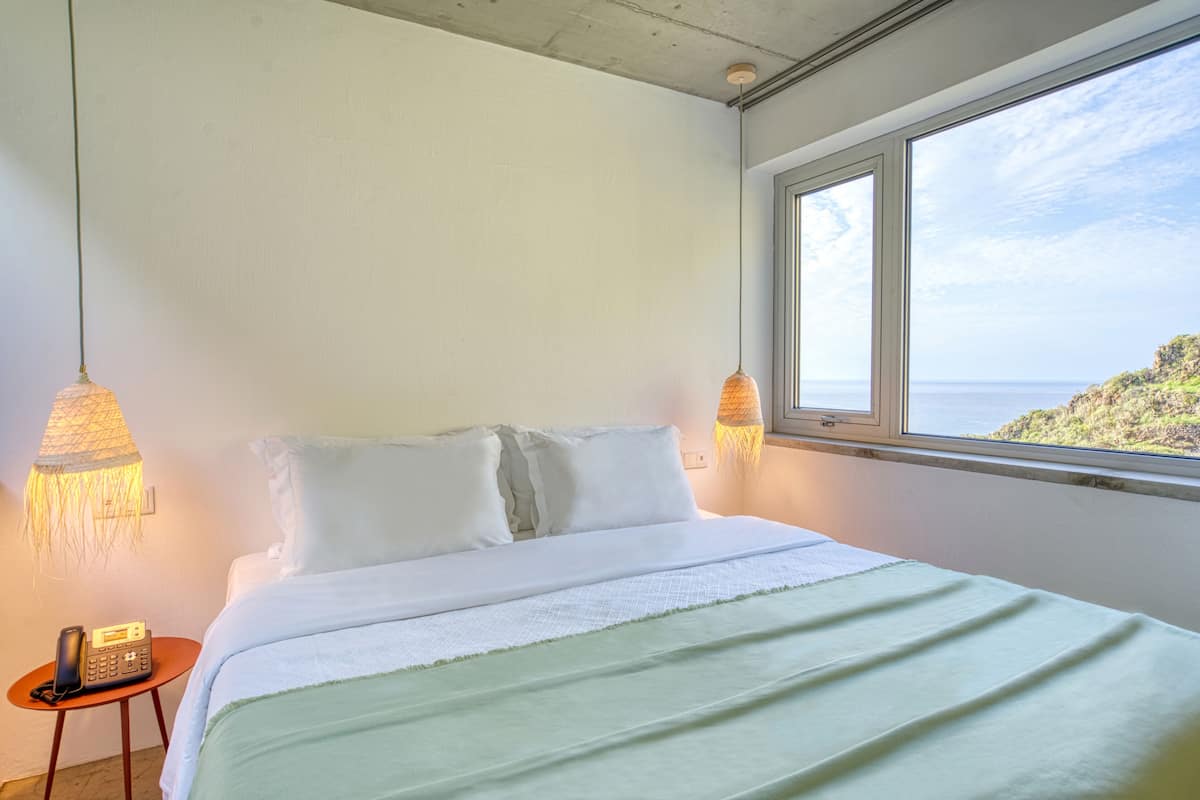 Quarto com cama, luminárias de cabeceira, telefone e janela com vista para um penhasco e o mar.