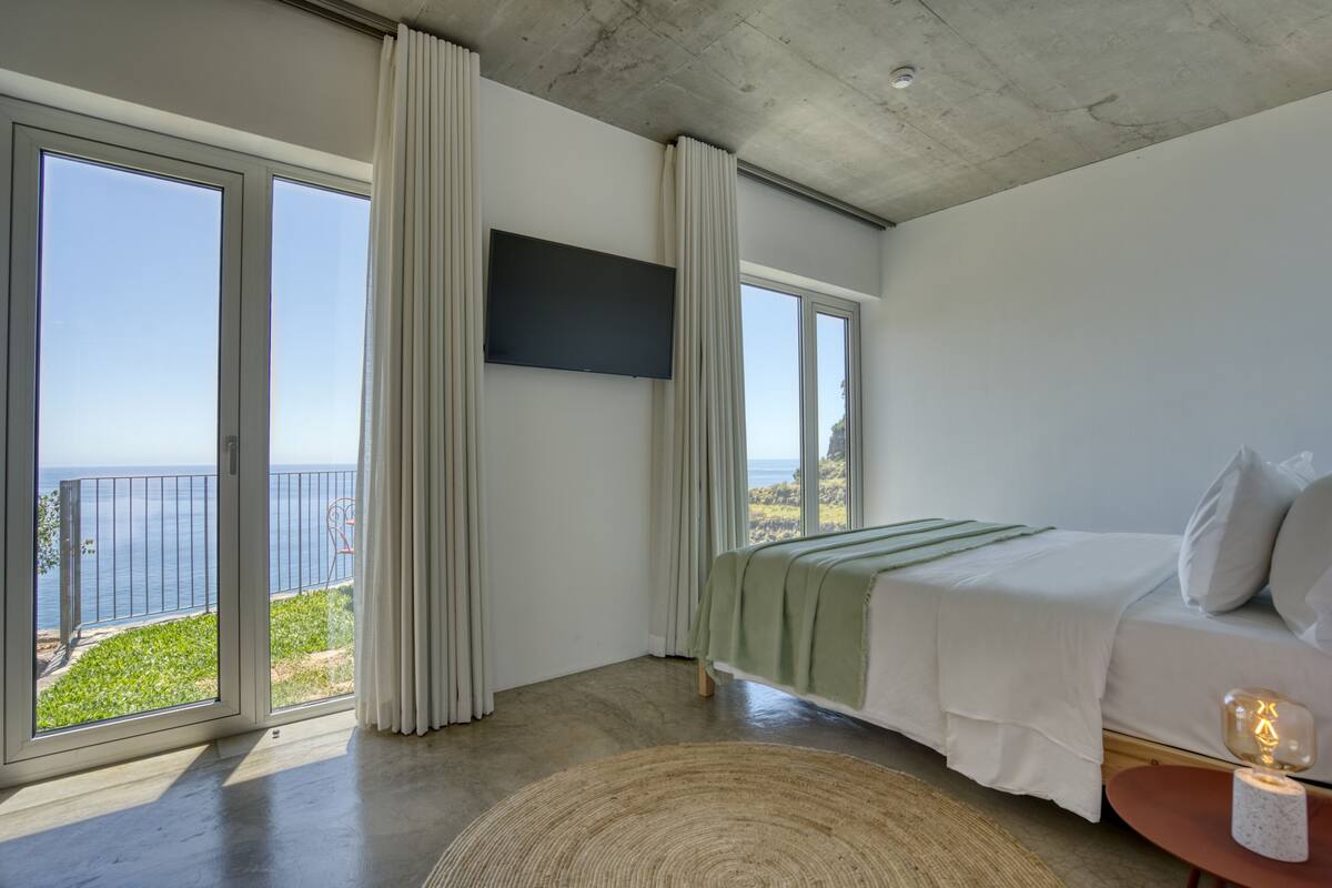 Quarto com cama grande, televisão de tela plana e vista para o mar através de grandes janelas.