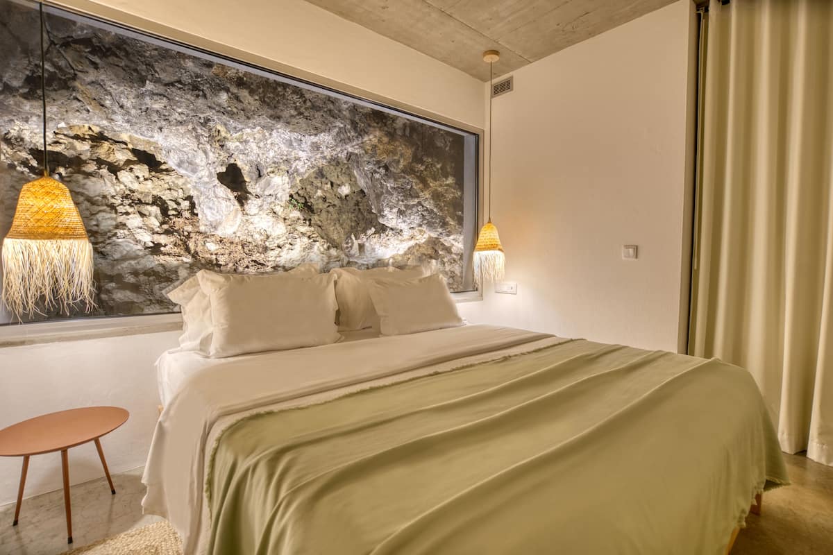 Quarto com uma cama grande, janela com vista para uma parede de rocha e duas luminárias pendentes.