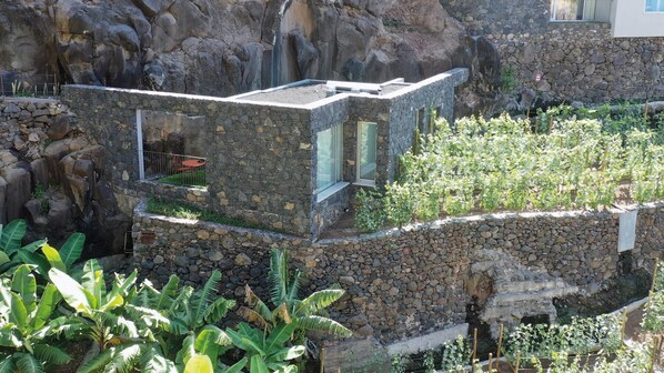 Edificio de piedra con techo plano, una ventana pequeña y un muro de piedra que rodea un jardín con diversas plantas.