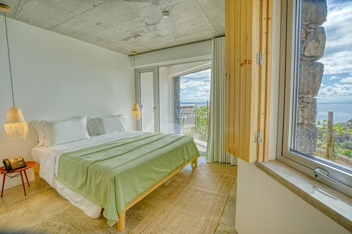 Quarto com cama, mesa de cabeceira, luminária e vista para o mar pela janela.