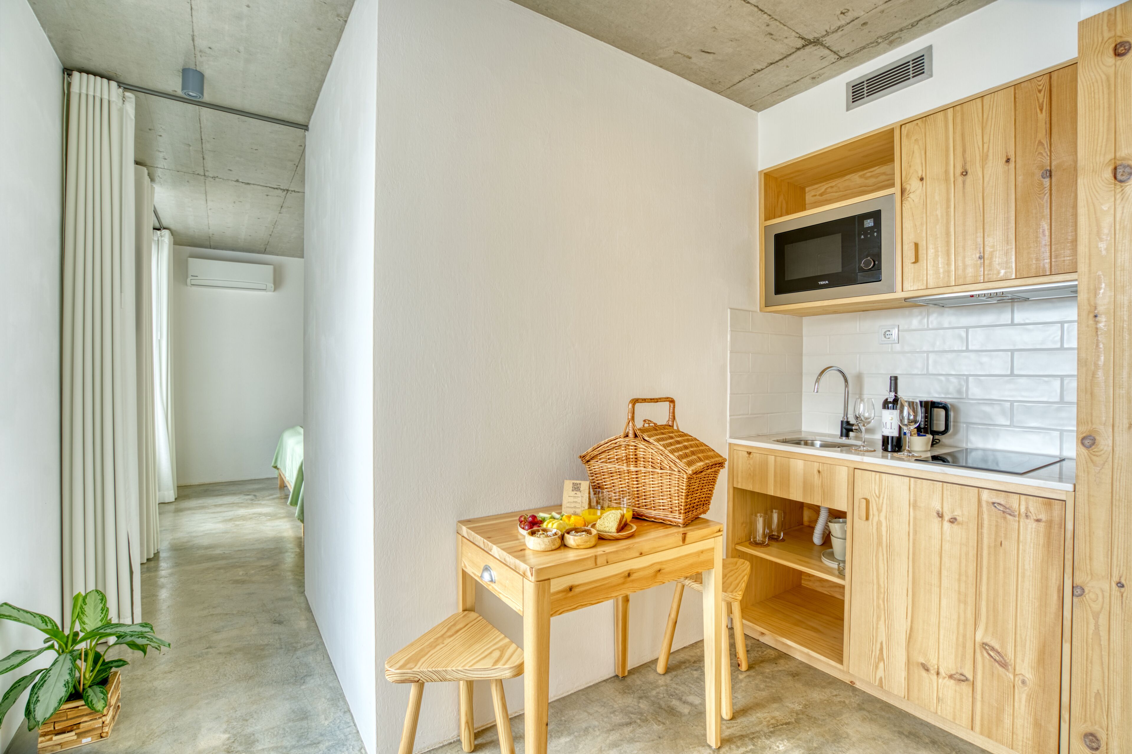 Cocina compacta con armarios de madera, microondas y un lavamanos. Hay una mesa de comedor con una cesta de fruta y una botella.