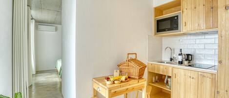 Cocina compacta con armarios de madera, microondas y un lavamanos. Hay una mesa de comedor con una cesta de fruta y una botella.