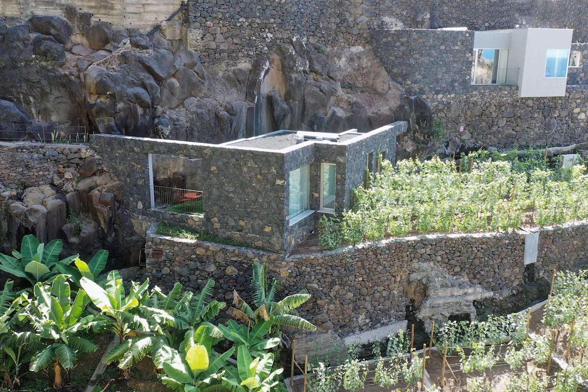 Um edifício de pedra com uma extensão moderna em vidro, cercado por vegetação exuberante e uma paisagem rochosa.