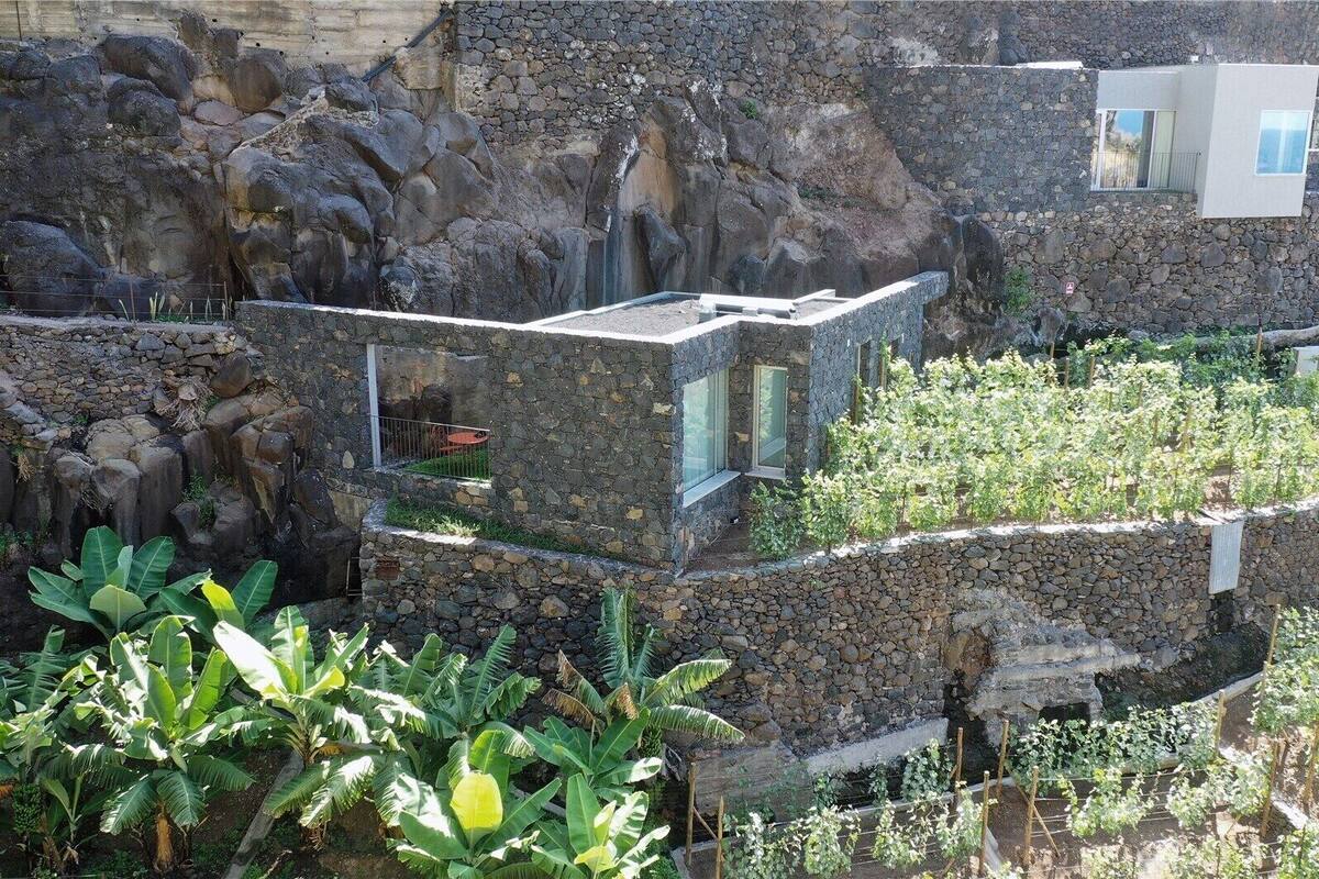 Edifício de pedra com extensão de vidro moderna, rodeado por vegetação exuberante e terreno rochoso.