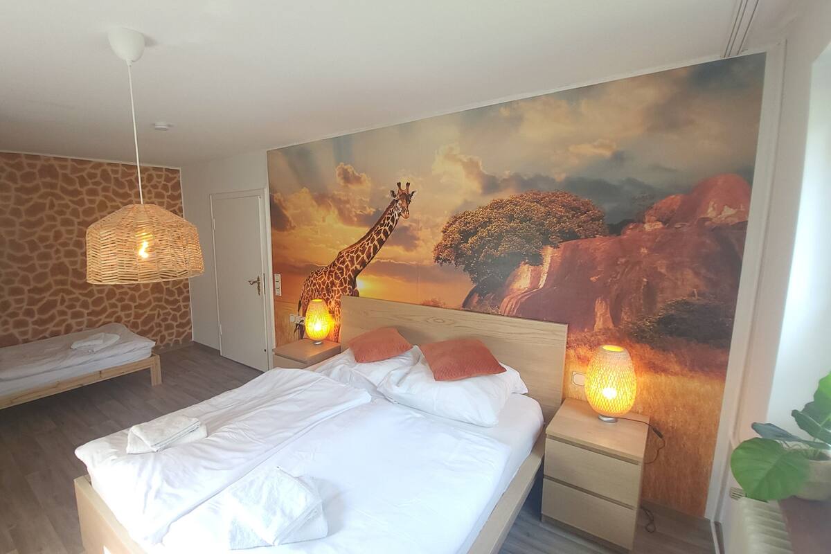 A hotel room with a bed, a bench, a wall mural of a giraffe, and a hanging light fixture.