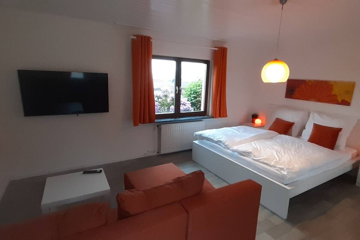 A modern bedroom with a bed, a TV, a window with curtains, and a bedside table with a lamp.