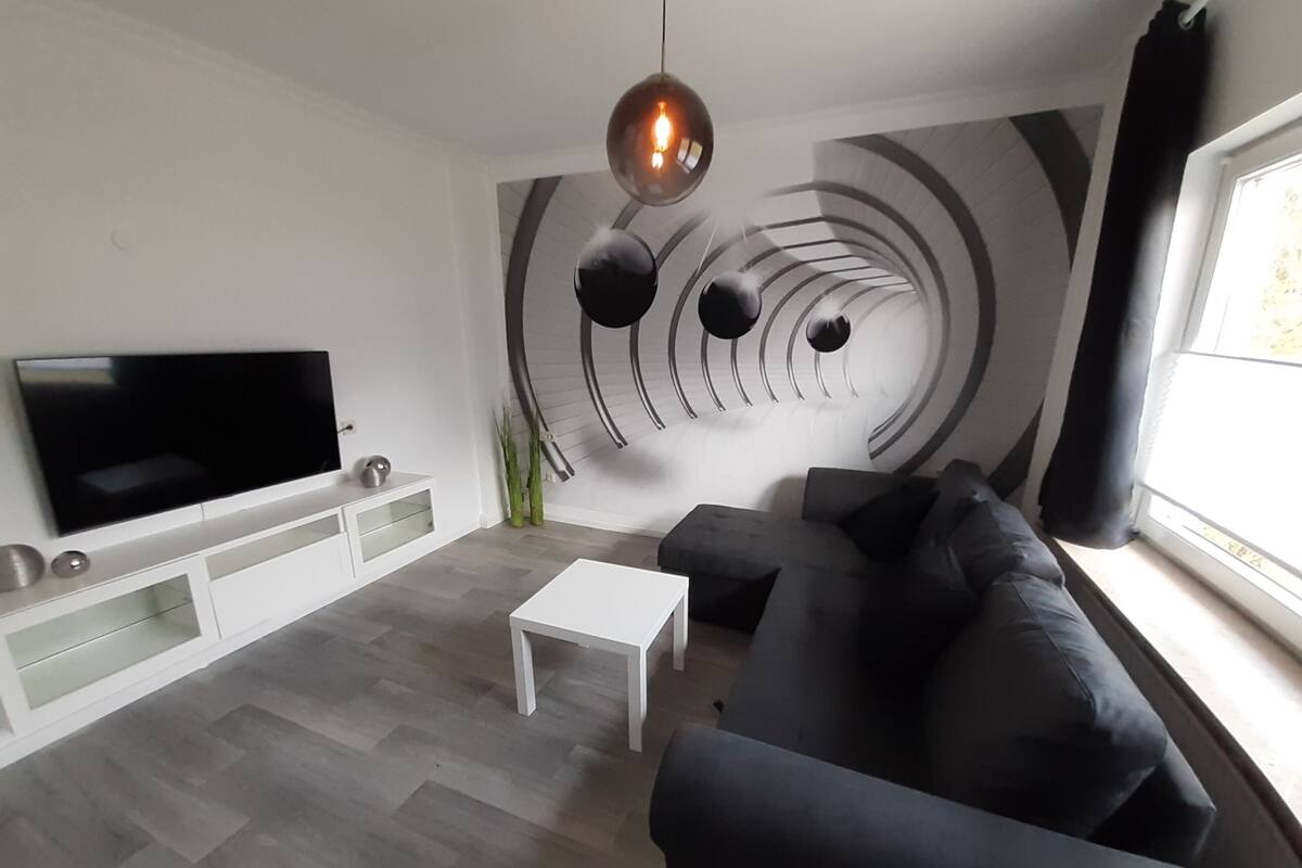 A modern living room with a flat-screen TV, a white coffee table, a black sofa, and a unique circular wall design.