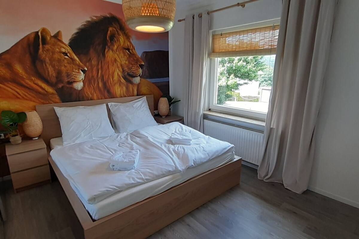 A bedroom with a large mural of lions, a wooden bed, white bedding, a bedside table with a lamp, and a window with curtains.