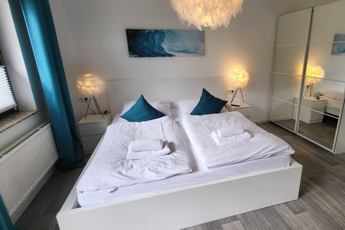 A modern bedroom with a large bed, two bedside tables with lamps, a large mirror, and a view of greenery outside.