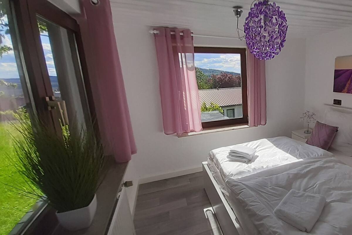 A bedroom with a bed, a window with curtains, a nightstand, a wall-mounted painting, and a potted plant.