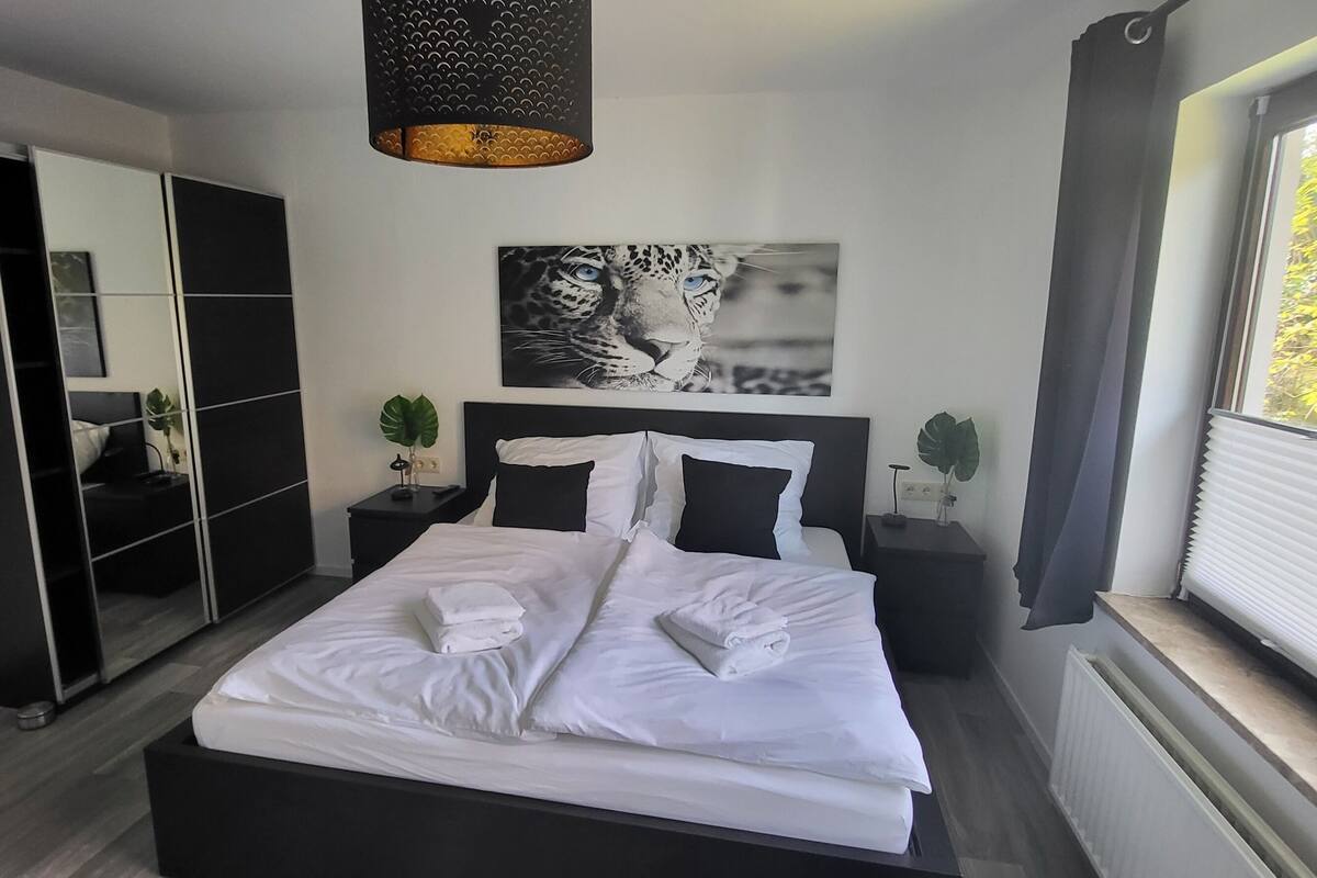 A modern bedroom with a large bed, a wardrobe, a mirror, a painting of a leopard, and a window with blinds.