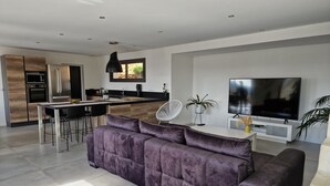 A modern living room with a large purple sofa, a dining area with a kitchen in the background, and a flat-screen TV on a white stand. - Quiet modern villa with swimming pool (Le Tignet)