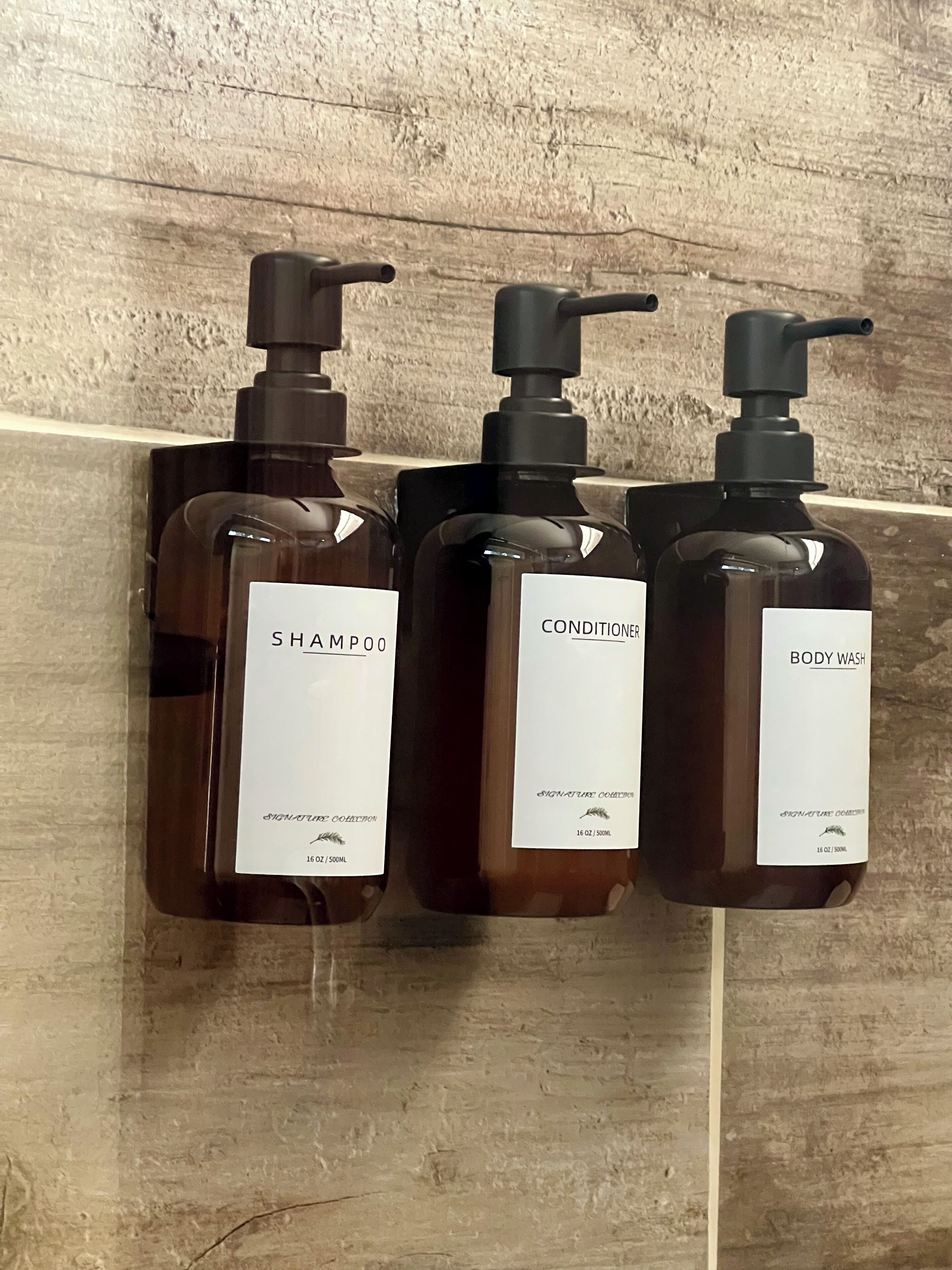Tre flasker fra Signature Collection: shampoo, balsam og body wash, hver med pumpe, monteret på en struktureret væg.