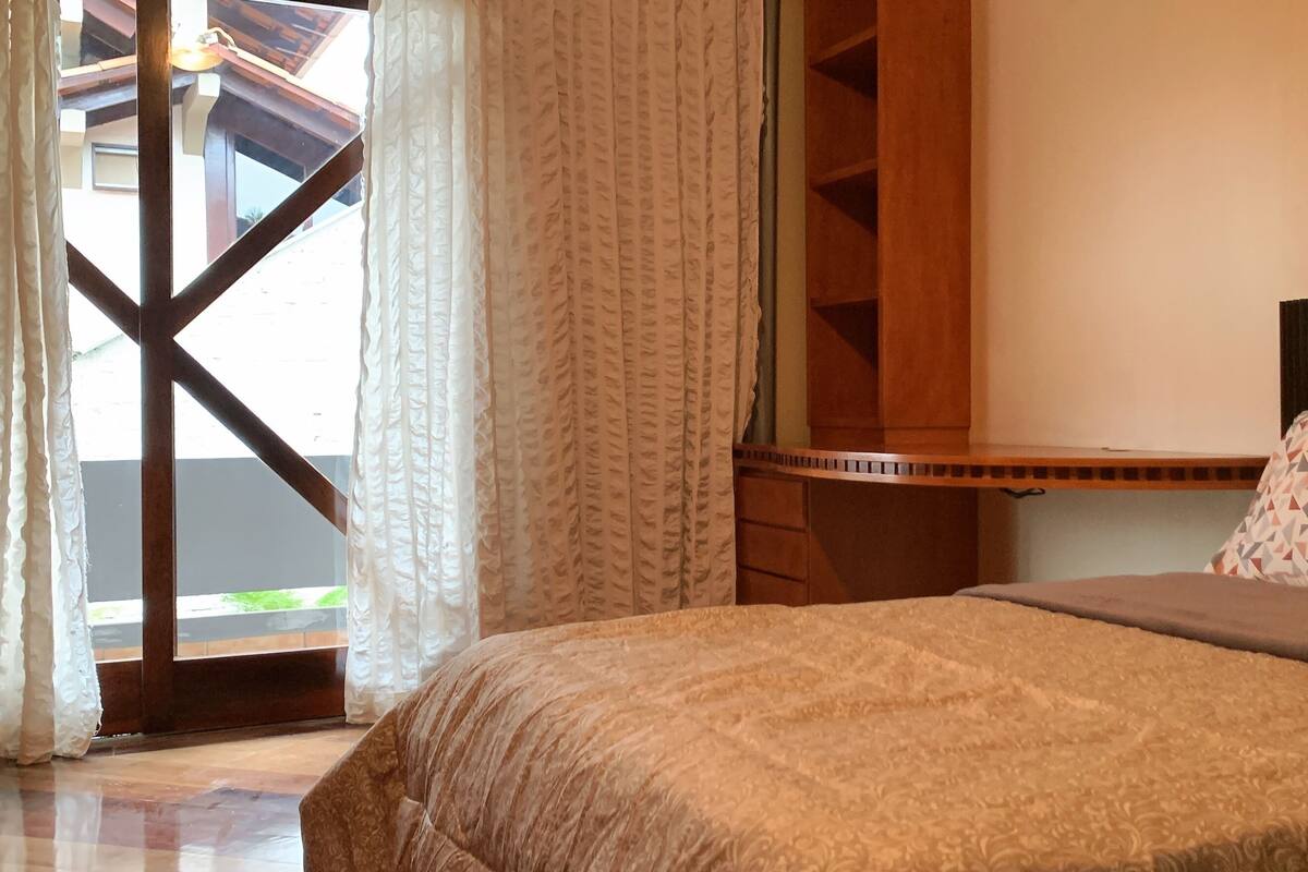 Un dormitorio con cama, almohadas estampadas, cabecera de madera y vista a través de una ventana con cortinas.