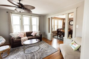 Ein geräumiges Wohnzimmer mit einem braunen Sofa, einem runden Couchtisch und einem Teppich mit Muster. Es gibt einen Deckenventilator, einen Essbereich mit Stühlen und eine Treppe, die in eine weitere Ebene führt.