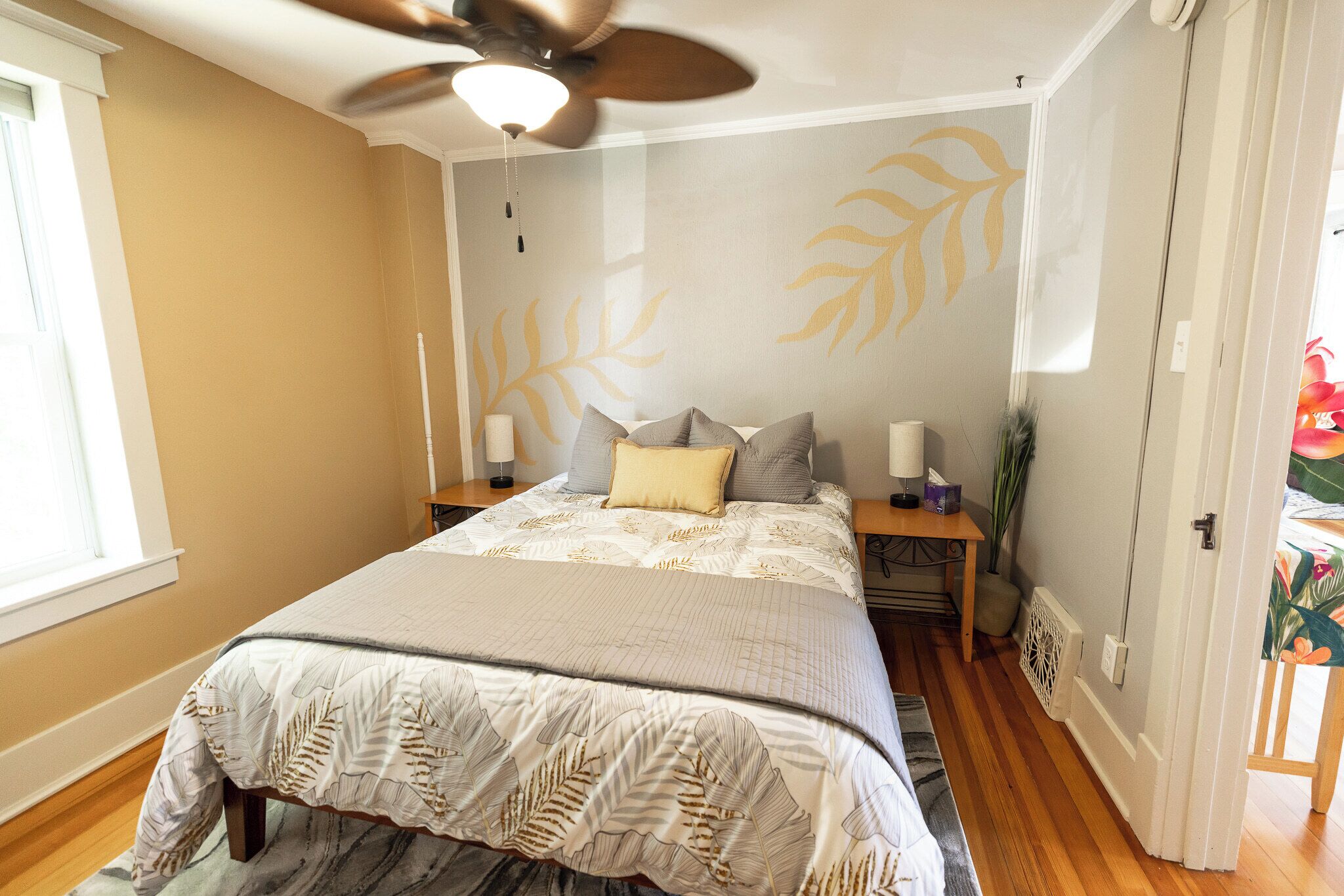 Ein Schlafzimmer mit einem Bett, einem Deckenventilator, einem Nachttisch, einer Lampe und einer Wand mit Blattmuster.