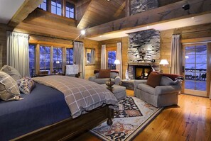 L’intérieur d’une cabane en rondins, avec un lit, deux fauteuils, une cheminée et de grandes fenêtres.