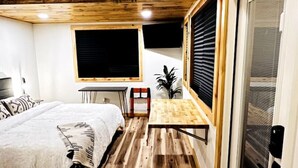 Un dormitorio moderno con techo de madera, una cama con manta blanca, una mesita pequeña y un ventanal grande con persianas.