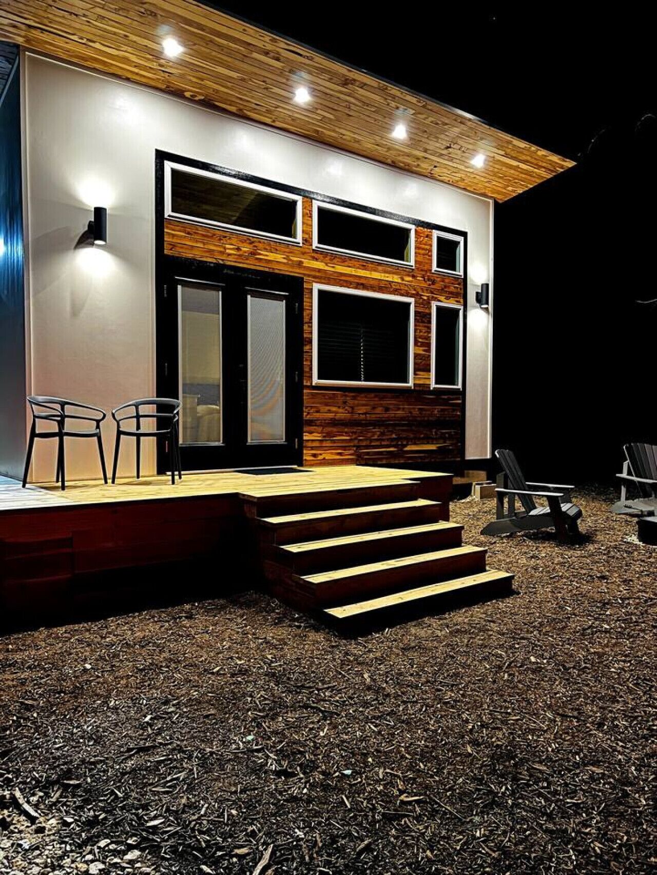 A modern building with a wooden facade, large windows, and a set of outdoor steps leading to a seating area with chairs.