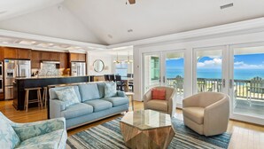 A modern living room with a sofa, armchairs, and a coffee table. There is a kitchen area with a refrigerator and a dining table. The room has large windows with a view of the ocean. - Seaside Oasis *Oceanfront Luxury w/ PRIVATE Beach Walkway, Pool, Elevator, Media Rm, Pool Table... (Nags Head)
