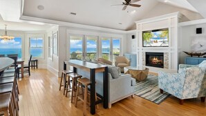 A spacious living room with a large window overlooking the ocean, a fireplace, and a cozy seating area. - Seaside Oasis *Oceanfront Luxury w/ PRIVATE Beach Walkway, Pool, Elevator, Media Rm, Pool Table... (Nags Head)