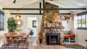 A cozy living room with a stone fireplace, a dining area with a wooden table and chairs, and a view of the kitchen in the background.